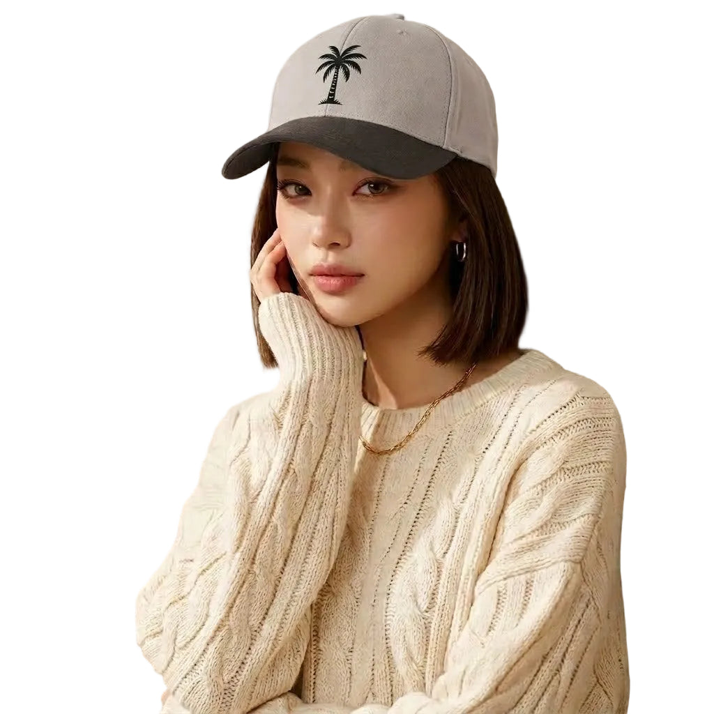Woman wearing a gray cap with a palm tree design, sitting against a textured wall with plants.