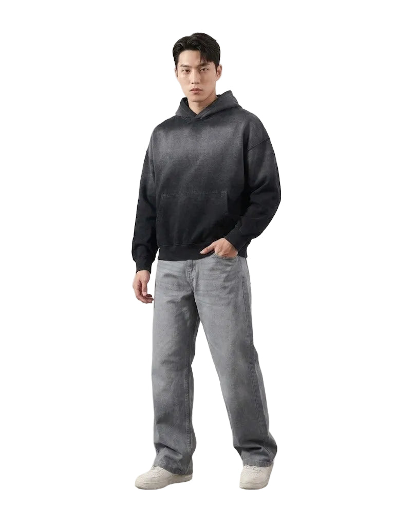Man wearing a dark hoodie and gray pants standing against a white wall.
